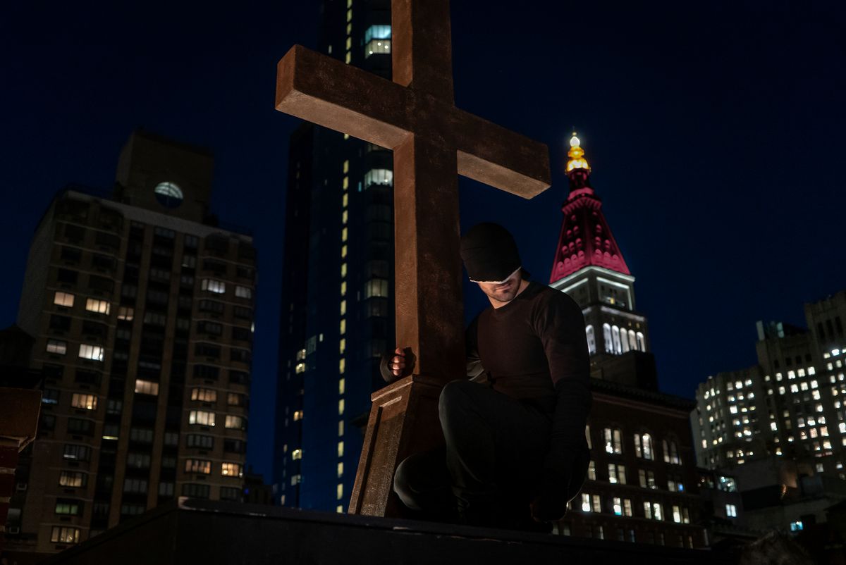 Daredevil in his black suit gripping a crucifix in front of the empire state building