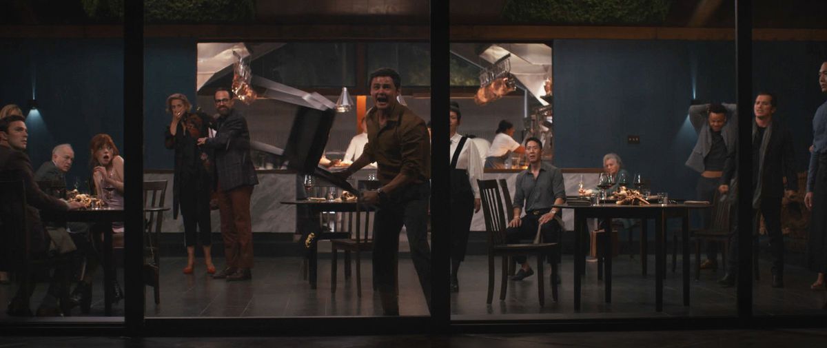 Un hombre frenético balancea una silla contra una de las paredes de vidrio de un elegante restaurante en un intento de romperla y escapar, mientras el resto de los clientes asustados observan una escena de El menú.