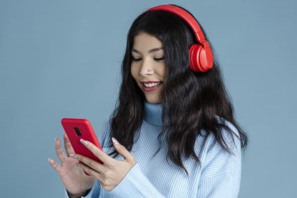 Una mujer con audífonos rojos y suéter azul sonríe a su teléfono inteligente mientras programa publicaciones en Instagram.