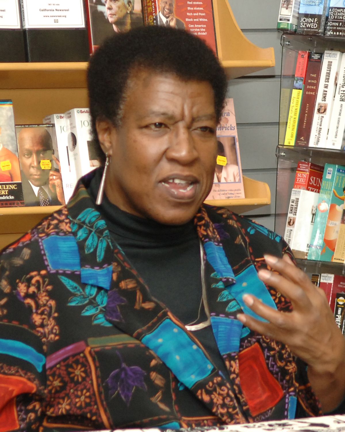 Octavia E. Butler in the 1990s, wearing a patterned shirt with tree leaf designs and long metal earrings, is caught mid-sentence during a reading at a book store