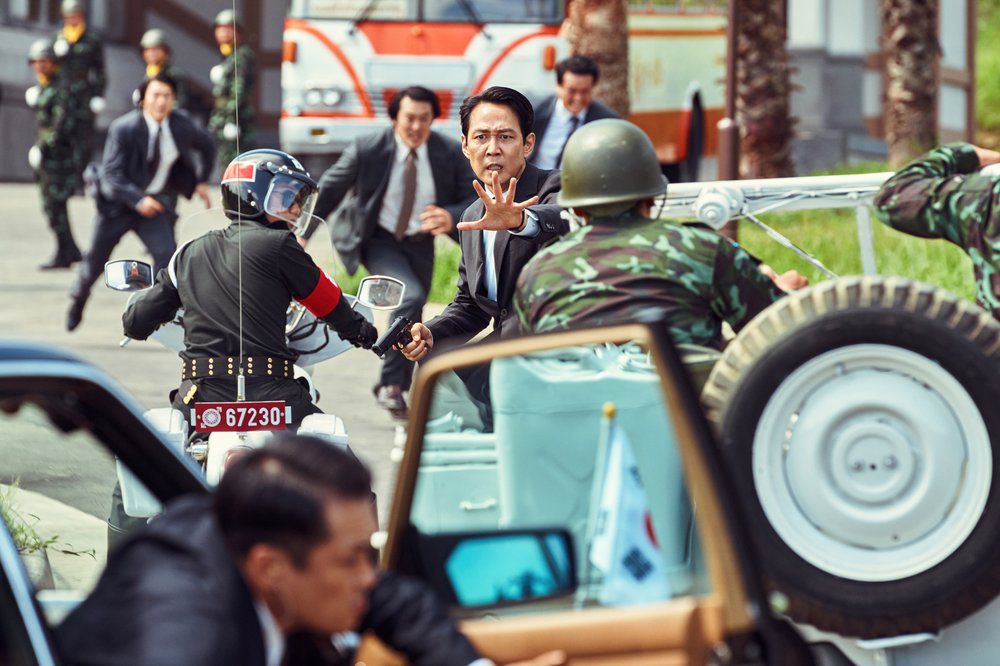 Lee Jung-jae tend la main dans une rue animée entourée de gens qui courent en costume et en tenue militaire à Hunt.