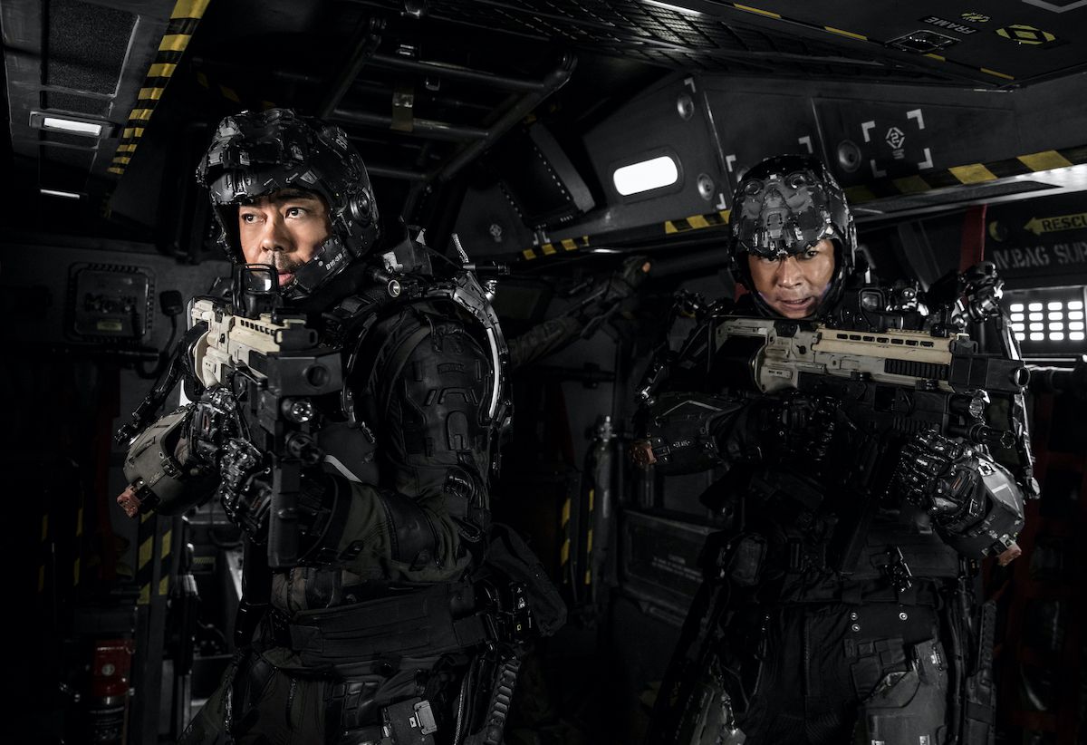 Hai người đàn ông mặc bộ đồ đen mang theo súng trường trong một hành lang công nghiệp khoa học viễn tưởng.