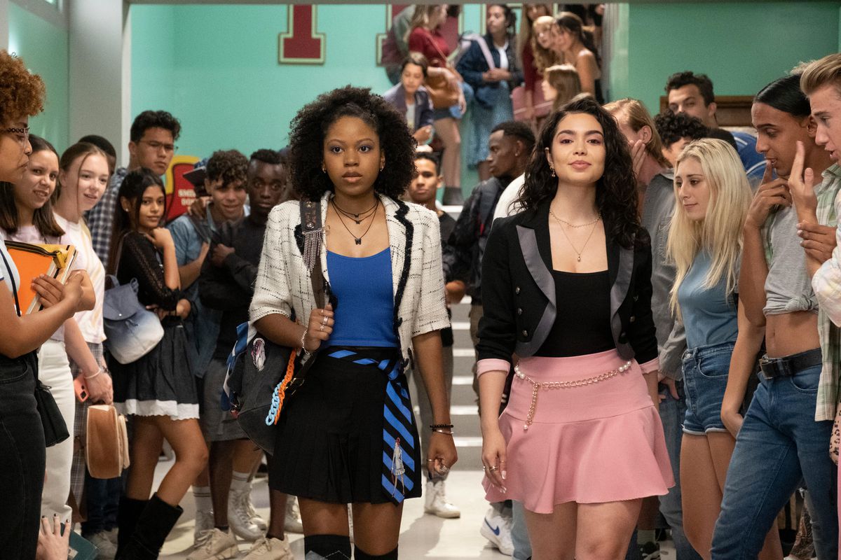 A woman in a black and blue outfit (Riele Downs) and a woman in a black and pink outfit (Auli’i Cravalho) walk down a hallway as a crowd of students look on around them.