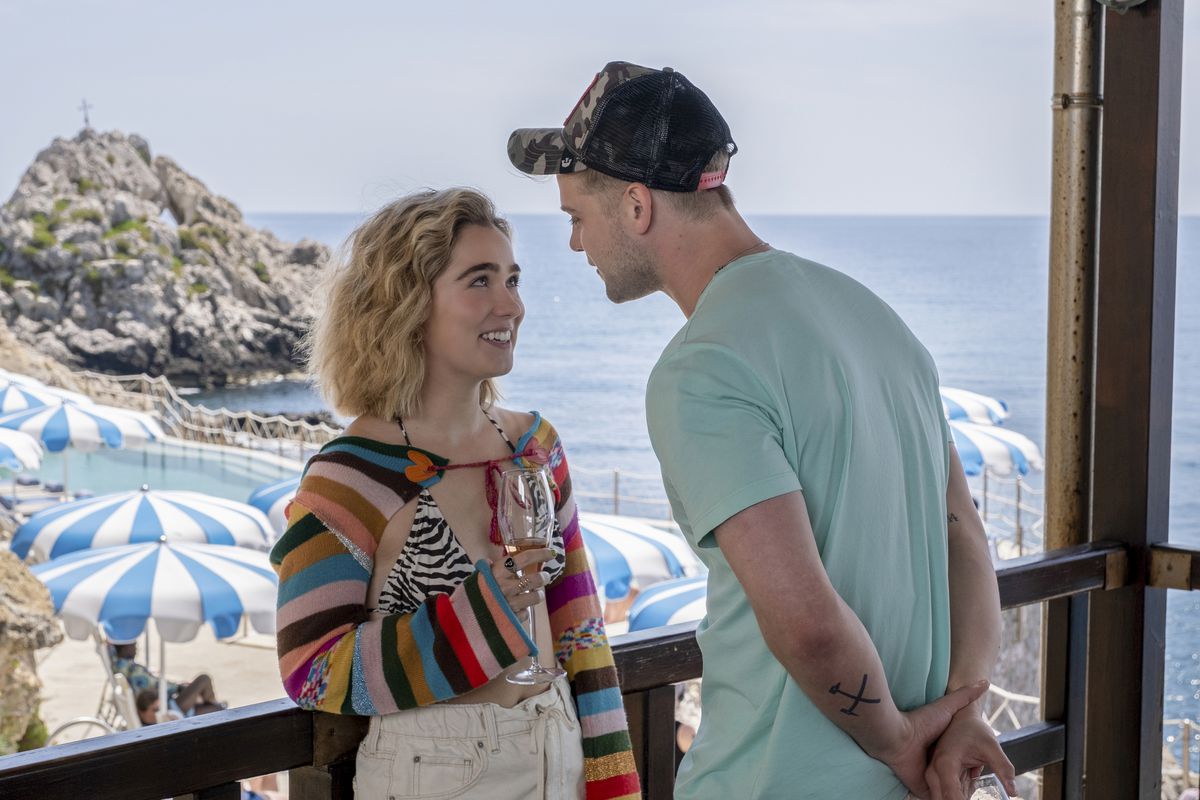 Haley Lu Richardson as Portia in White Lotus. She’s talking to a young man outside on a patio bar overlooking the ocean. She’s wearing a rainbow knit bolero top with a zebra-print bikini top.