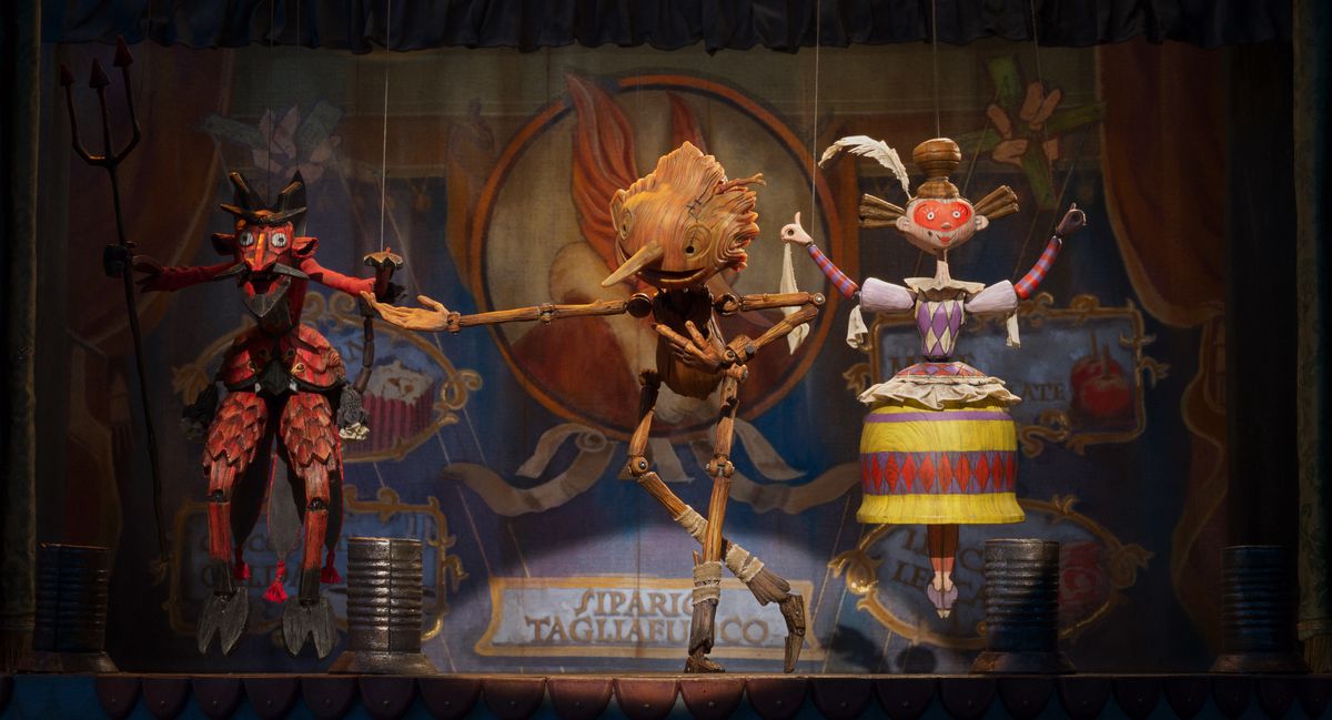 A wooden puppet boy bows on-stage beside two puppets painted as a demon and a woman in a costume.