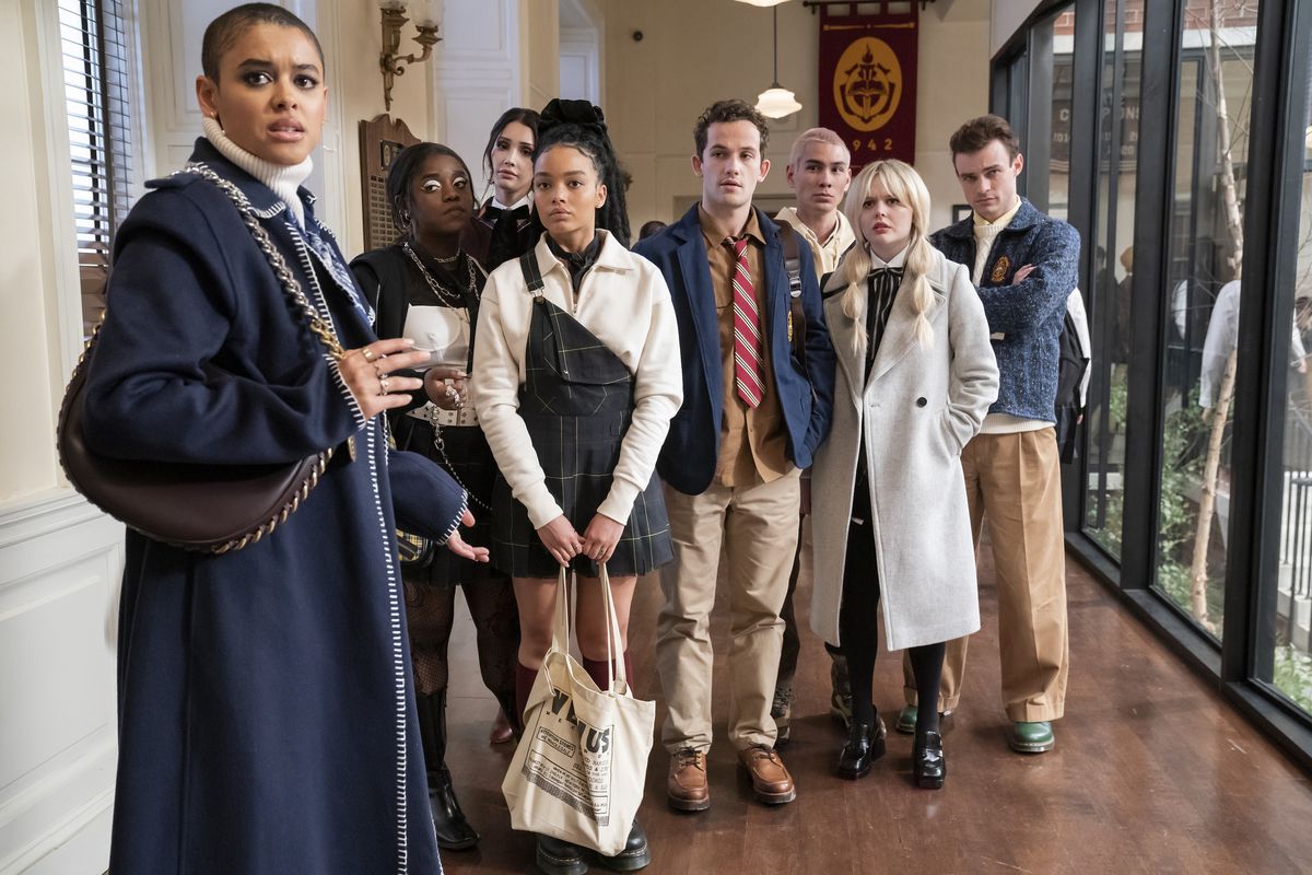 The main cast of Gossip Girl, all cloaked in their most fashionable fall attire, stand in the middle of their high school hallway facing the camera with concerned looks on their faces. 