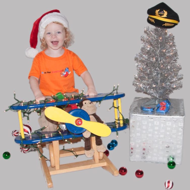 Een jong kind met een kerstmuts op een hobbelpaard in een vliegtuig en poseert naast een kerstboom met daarop een kapiteinshoed.