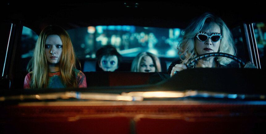 Jennifer Tilly drives a car, with a young person in the passenger seat and two dolls in the backseat, in Chucky.