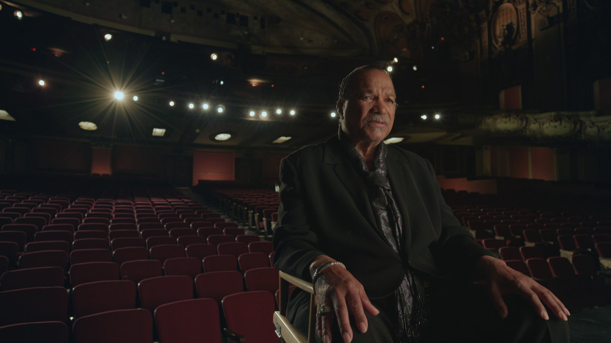 Billy Dee Williams zit in een regisseursstoel op een theaterpodium, met zijn rug naar de lege publieksstoelen gericht