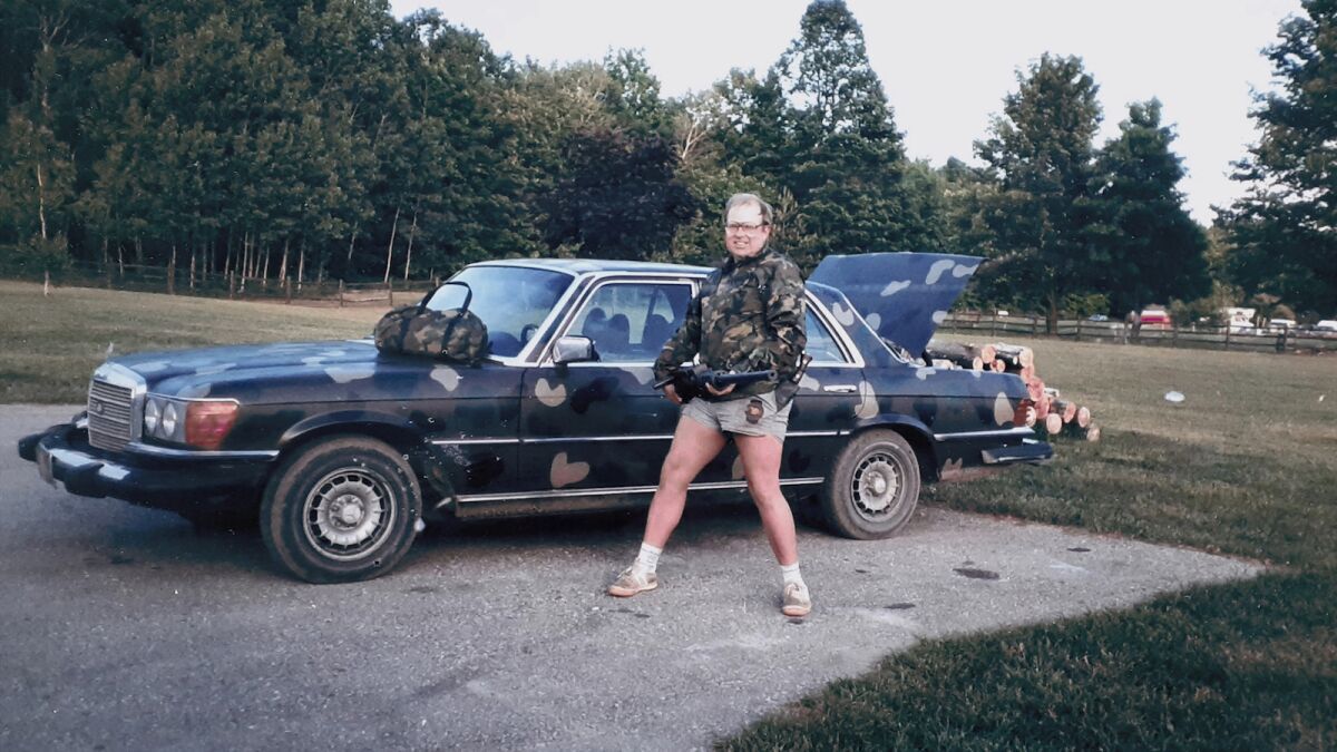 Een oude foto uit de jaren 1980 toont een man in camo en korte broek loopt over een veld naar een parkeerplaats waar zijn camo sedan geparkeerd staat
