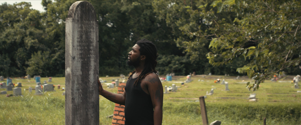 A Black man with dreadlocks and a black tanktop stands in a graveyard placing a hand on a towering gravestone inscribed with names