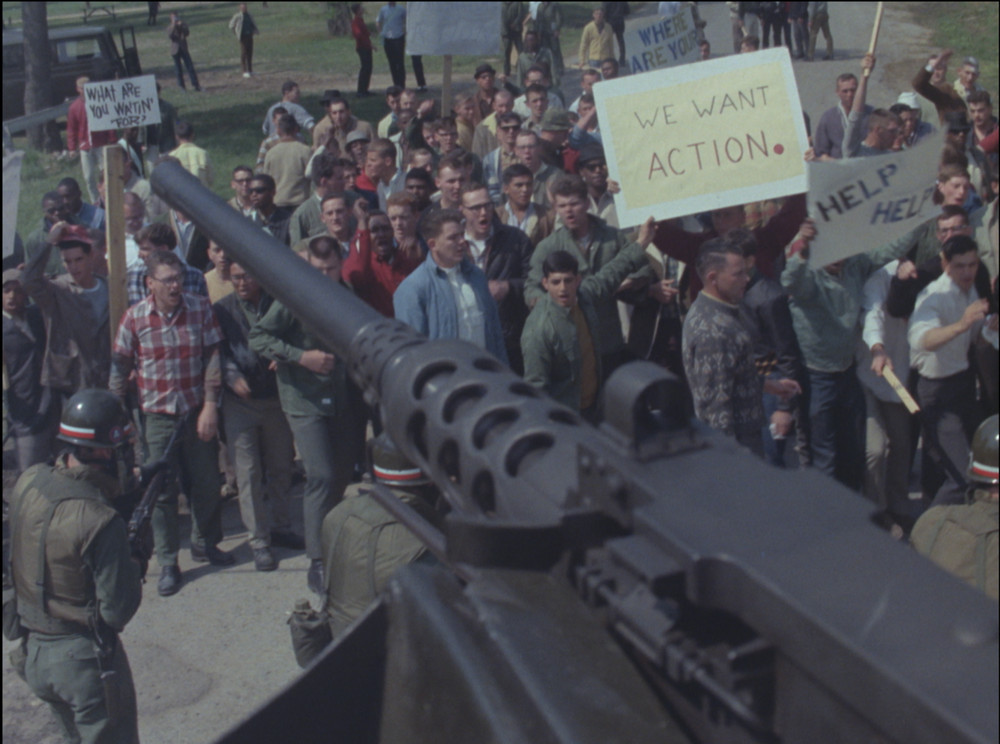 Een tankkanon springt in beeld boven een menigte opgevoerde demonstranten, van wie er één een bord vasthoudt met de tekst "We Want Action"