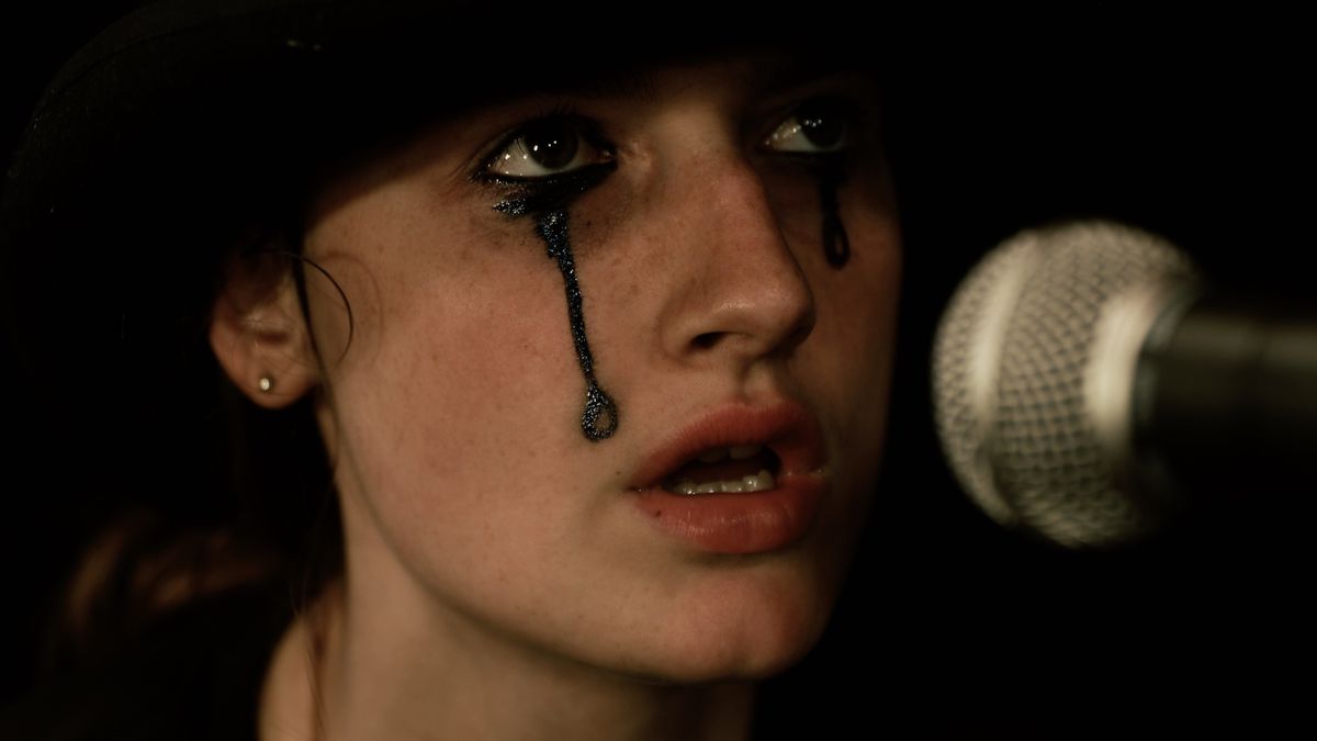زيلدا آدامز في دور إيزي في هيلبيندر ، تغني في ميكروفون بينما ترتدي قبعة سوداء مع مكياج أسود يركض من عينيها.