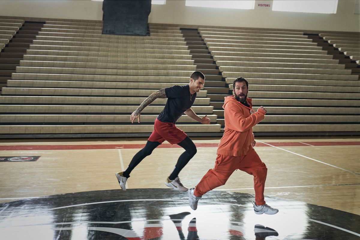 Adam Sandler, wearing an orange sweat suit, runs on a basketball court next to a smiling Juancho Hernangómez in Hustle.