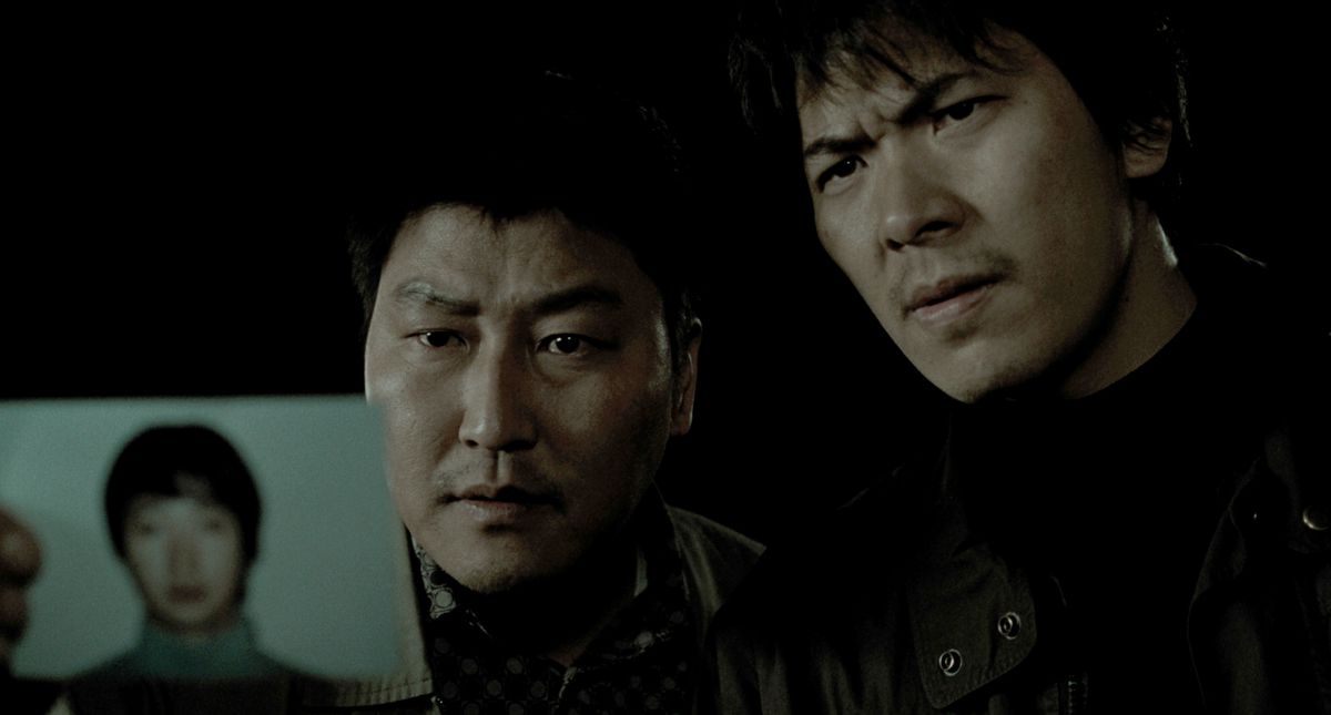 Dedektif Park (Song Kang-ho) ve Seo (Kim Sang-kyung), Memories of Murder'da cinayet zanlısı Park Hyeon-gyu'nun (Park Hae-il) fotoğrafını tutuyor.
