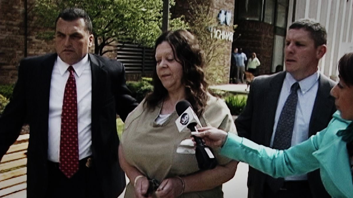 Một người phụ nữ mặc áo sơ mi màu nâu và bị còng tay bởi hai người đàn ông to lớn mặc com lê đang được phỏng vấn bởi một người phụ nữ mặc áo cộc tay màu ngọc lam đang cầm micrô.