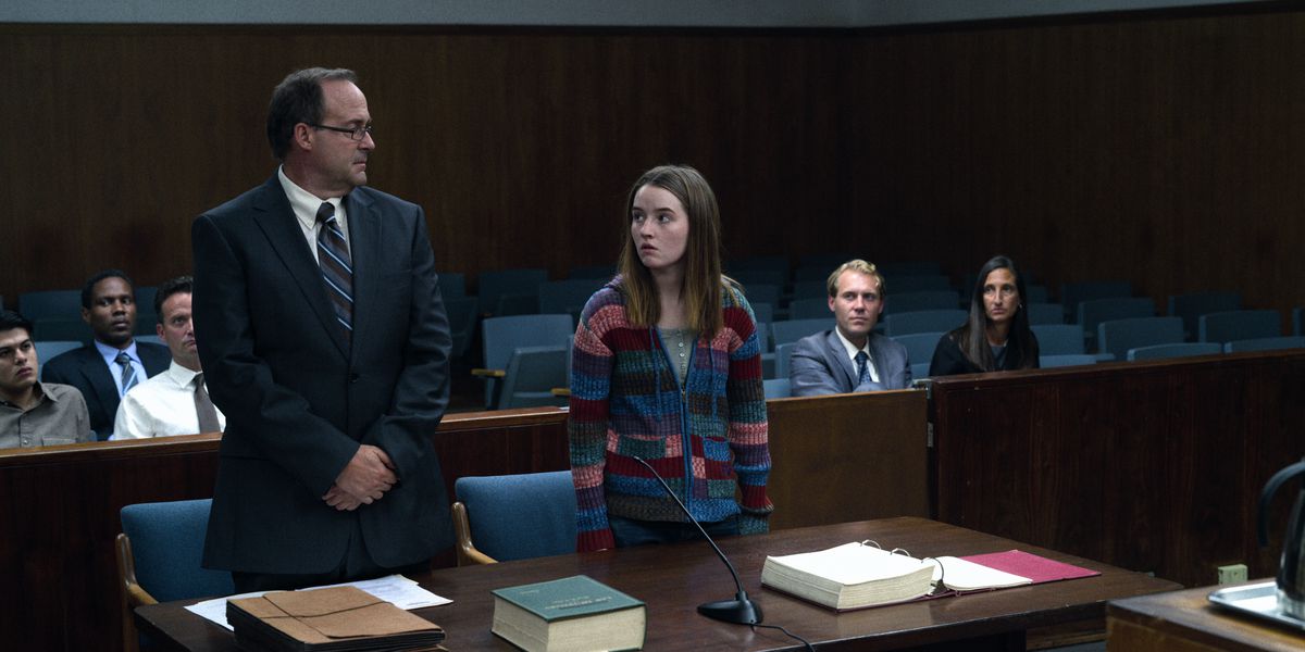 Çok renkli bir süveter giymiş genç bir kadın, mahkeme salonunda lacivert takım elbiseli bir adamın yanında duruyor.