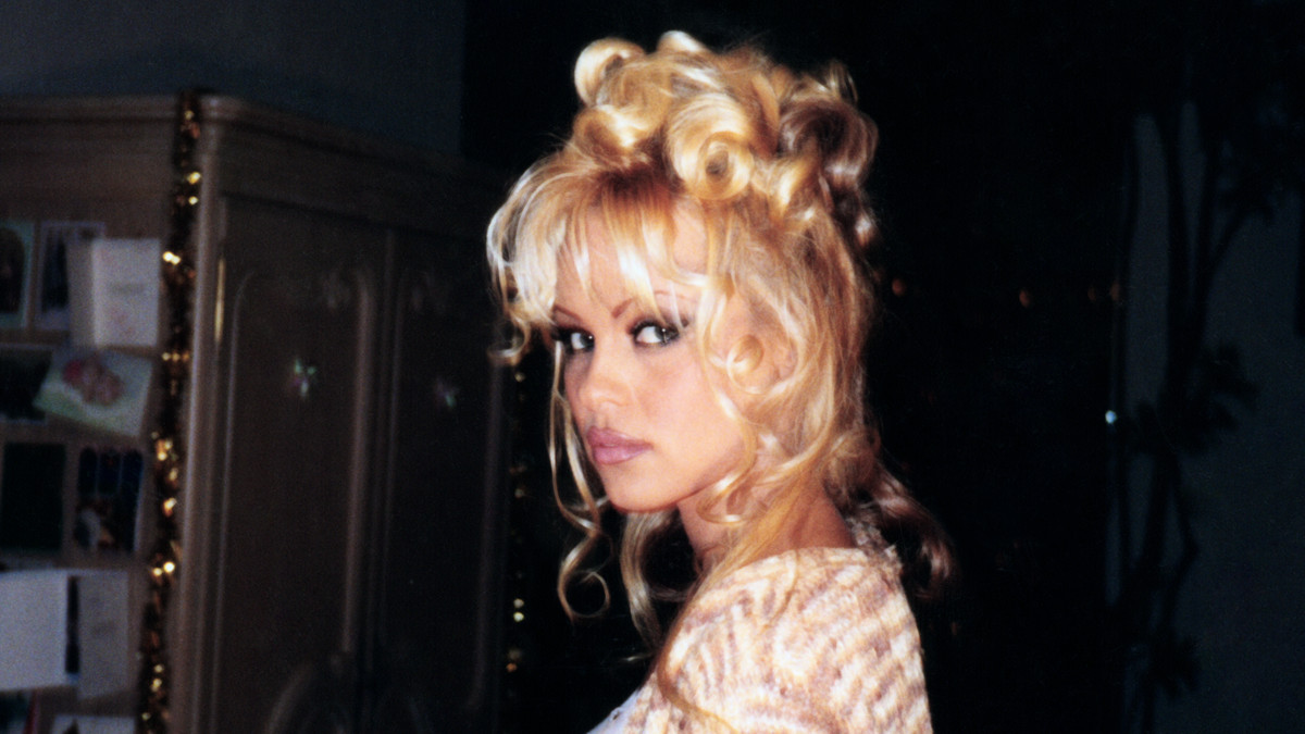 pamela anderson, peering at the camera. her blonde hair is piled on her head and her eyes are ringed with dark eyeliner. she is not smiling. 