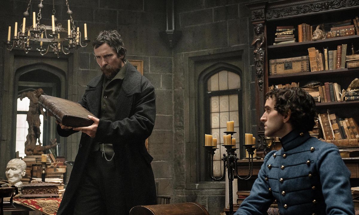 1830s detective August Landor (Christian Bale) grimaces at a book in a candle-filled West Point military academy library as cadet Edgar Allen Poe (Harry Melling) looks on in The Pale Blue Eye