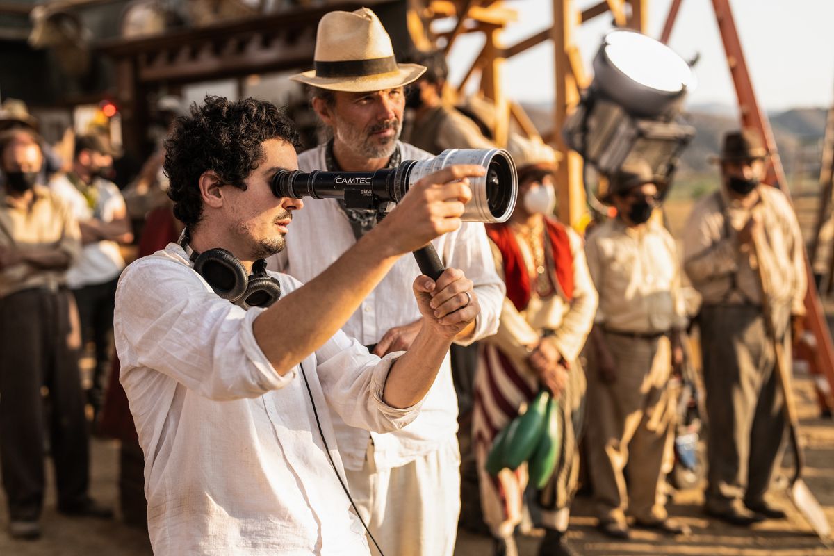 Damien Chazelle holds up a director viewfinder on the set of Babylon while DP Linus Sandgren stands behind him in a hat with a group of period-dressed extras stands in the background near a fake movie set