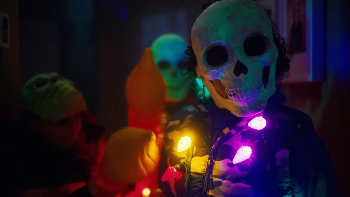 Kafatası suratlı bir maske ve boynuna çok renkli ışıklar saçılmış ayrıntılı bir kostüm giyen bir kişi.