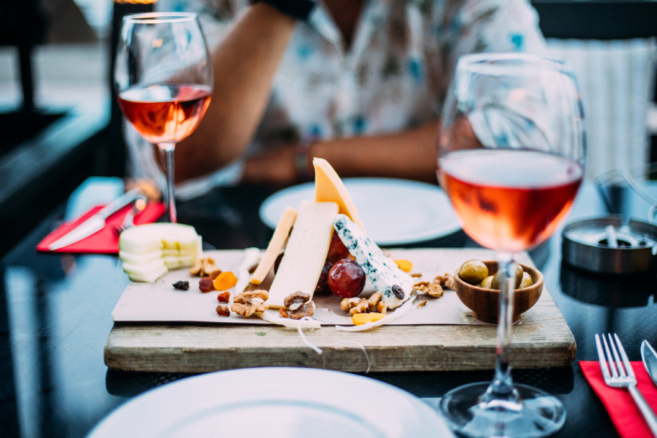 Dos copas de vino rosado y tablero con frutas, pan y queso en mesa de madera