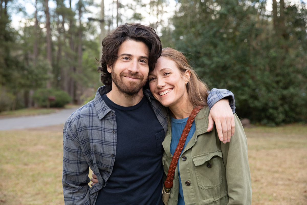 A man (Jake Hoffman) in a dark blue shirt poses for a photo with a smiling woman (Schuyler Fisk) in an olive green jacket.
