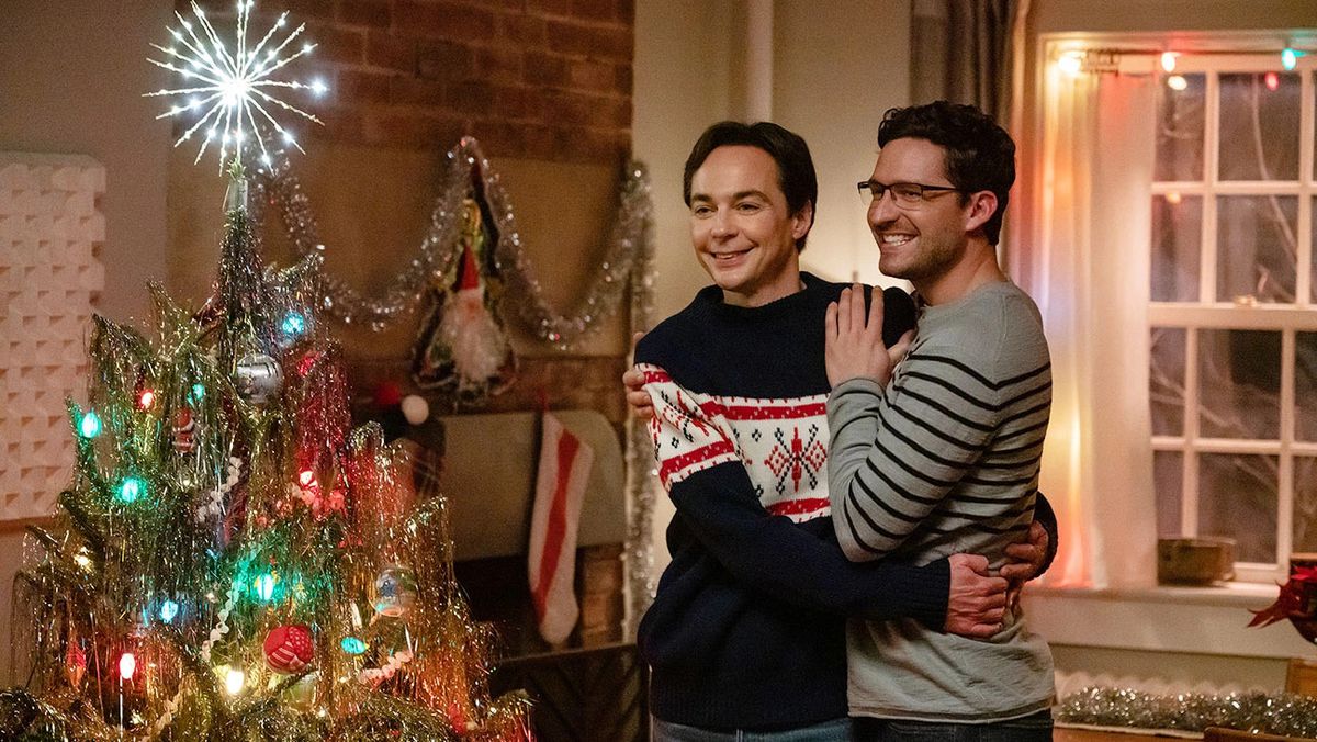 Two men (L-R: &nbsp;Jim Parsons, Ben Aldridge) in Christmas sweaters embrace one another as they stare lovingly at a Christmas tree together.