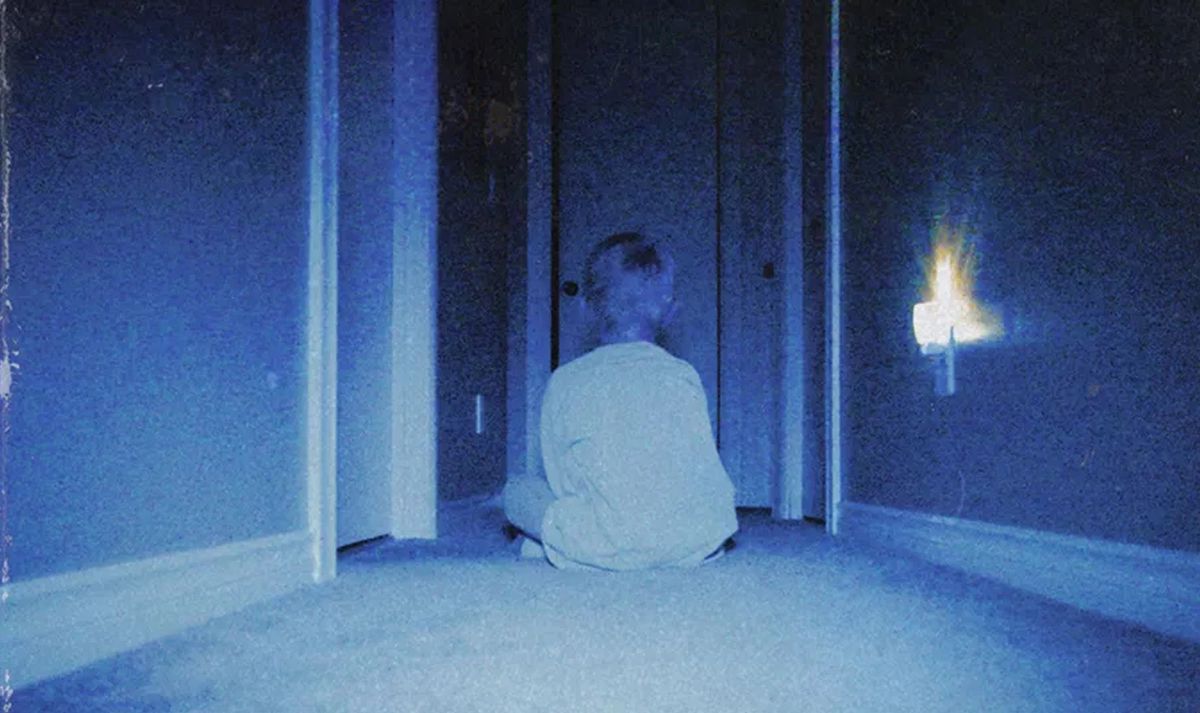 En ung pojke sitter i en mörk, blå korridor med ryggen mot kameran, vänd mot en serie öppna dörröppningar, i en typiskt kornig, luddig bild från skräckfilmen Skinamarink
