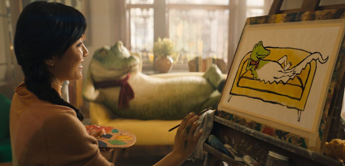 A dark-haired woman paints a cartoonish portrait of a crocodile lounging on a yellow couch with a scarf wrapped around its neck.