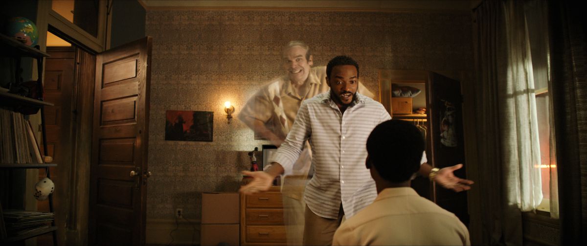 A man (Anthony Mackie) speaks to his son sitting on a bed with his hands raised with a smiling ghost (David Harbour) peering over his shoulder.