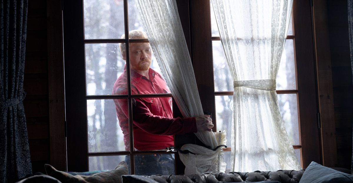 Redmond (Rupert Grint), a slight, red-haired man in a bright red shirt, breaks through the glass of a set of patio doors while forcing an entrance into the cabin in Knock at the Cabin