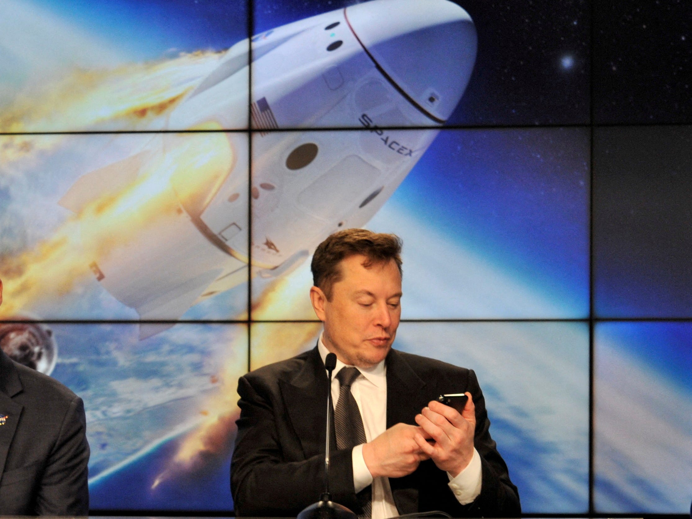 Elon Musk mira su teléfono con una pantalla de una cápsula espacial entrando en órbita detrás de él.