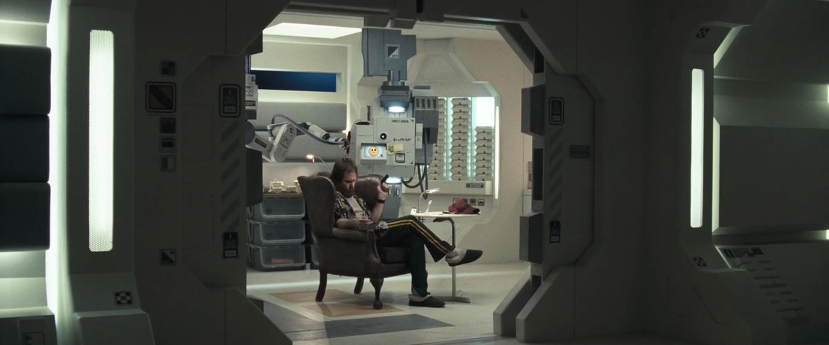 Een man met een baard in een kamerjas en pantoffels (Sam Rockwell) zit in een bruine leunstoel in een futuristische gang met een lachend computerscherm over zijn schouder.