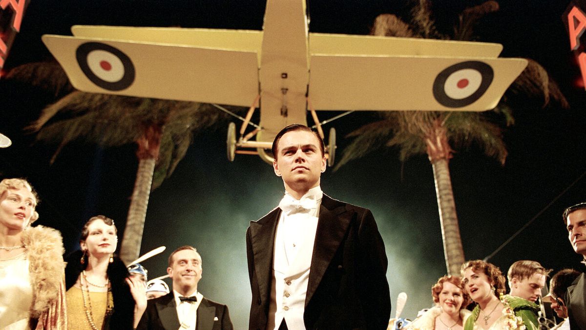 Một người đàn ông trong bộ tuxedo (Leonardo DiCaprio) đứng bên dưới chiếc máy bay hai tầng màu vàng lơ lửng phía trên anh ta trong khi hai bên là những hàng người xem ăn mặc lộng lẫy và những cây cọ.
