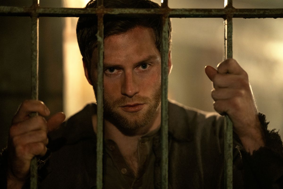 Matthias, a handsome, broad, blonde man, presses his face against the bars of a jail cell