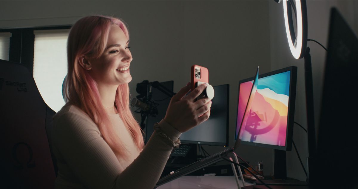 Eine rosahaarige Frau (Siri Dahl), die an einem Schreibtisch sitzt, lächelt, während sie eine Handykamera hält