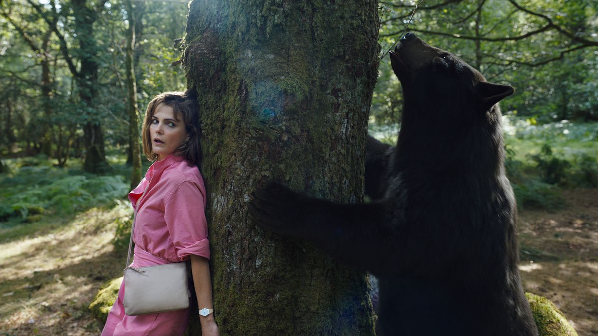 Sari (Keri Russell), eine verängstigt aussehende Frau in einem rosa Kleid, versteckt sich hinter einem Baum, während ein von Kokain benebelter Bär in Cocaine Bear auf der anderen Seite herumschnüffelt
