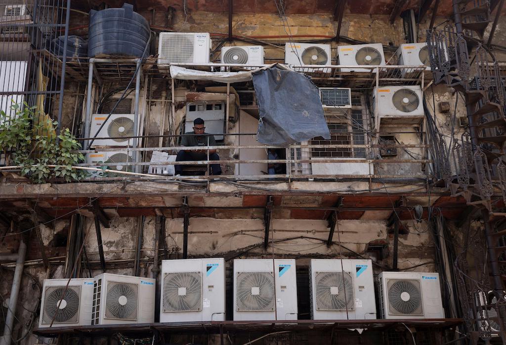 Een man gebruikt zijn mobiele telefoon terwijl hij tussen de buitenste units van airconditioners zit, aan de achterkant van een commercieel gebouw in New Delhi, India op 30 april 2022.