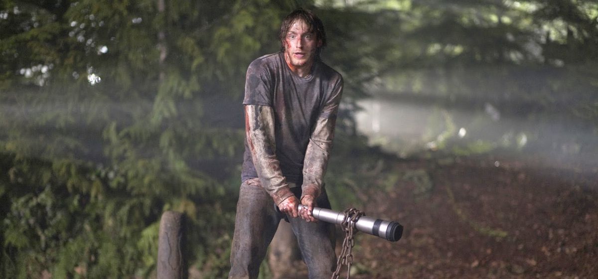 Marty (Fran Kranz), a stoner in filthy jeans and a grey T-shirt covered in muck and blood, stands in the woods holding a telescoping silver bong as a club with a rusty chain wrapped around it in a scene from 2011’s The Cabin in the Woods