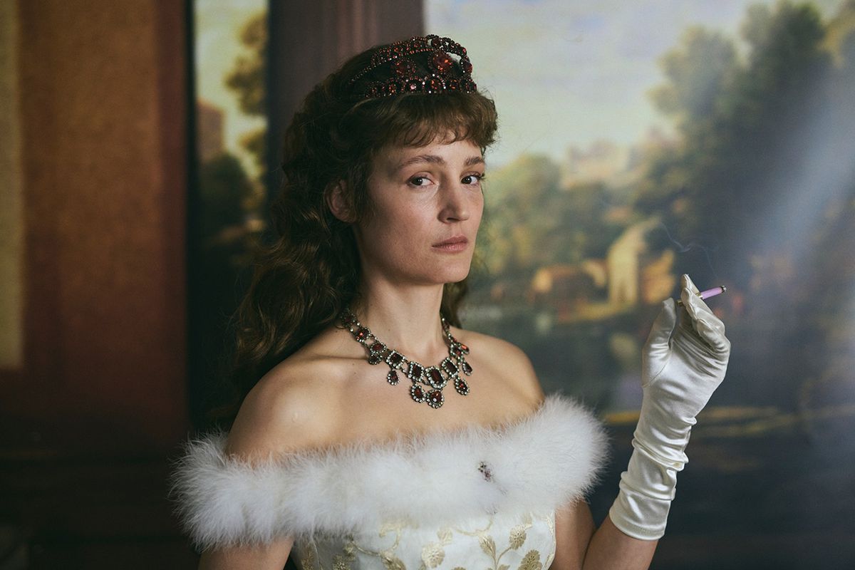 Vicky Krieps sigara içiyor ve Korsaj'da Avusturya İmparatoriçesi Elisabeth olarak süslü bir tablonun önünde bir taç, süslü bir elbise ve beyaz bir eldiven giyiyor.
