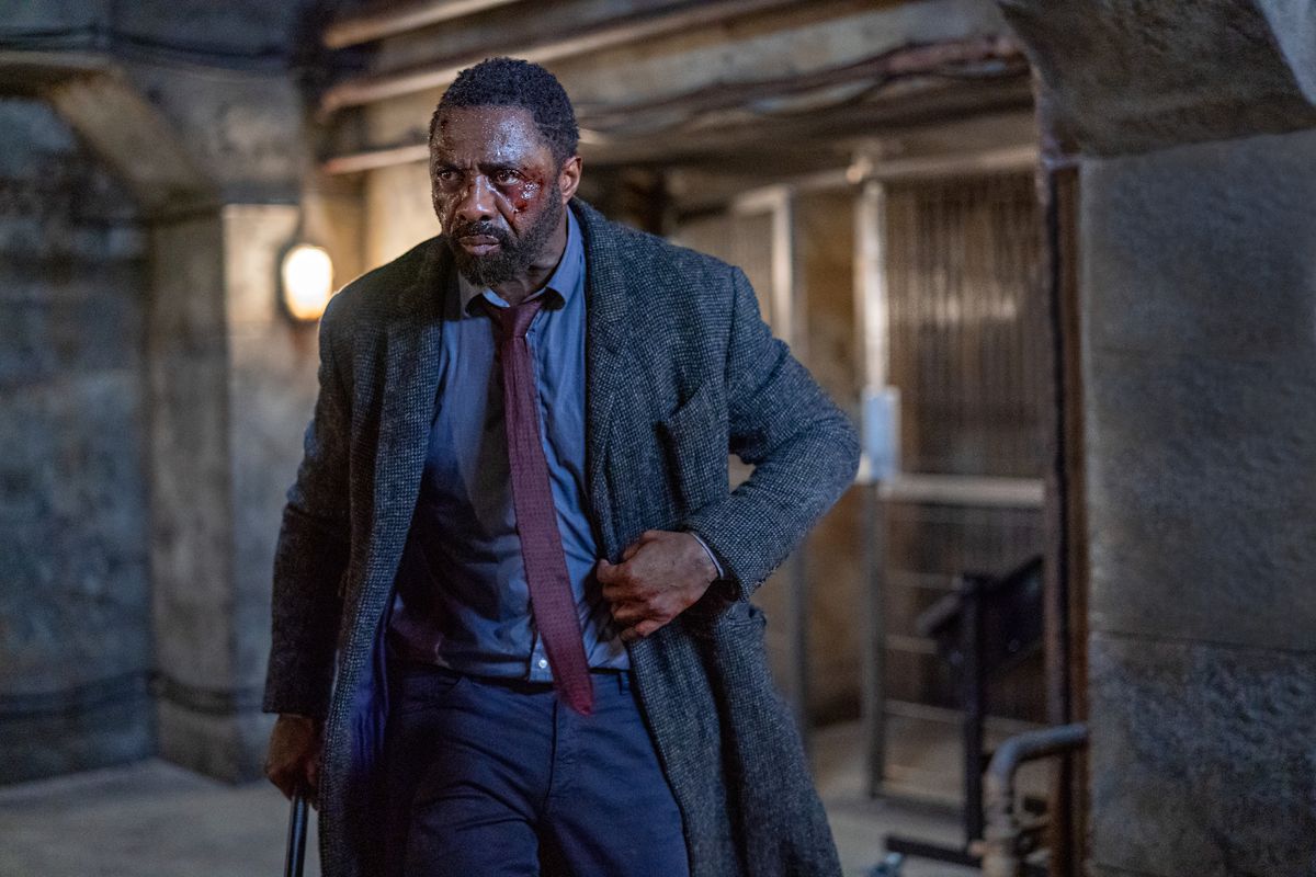 Ένας άντρας (Idris Elba) ντυμένος με ένα μπλε πουκάμισο, μπορντό γραβάτα και ένα μακρύ παλτό τουίντ με εμφανείς μώλωπες και αίμα στο πρόσωπό του.