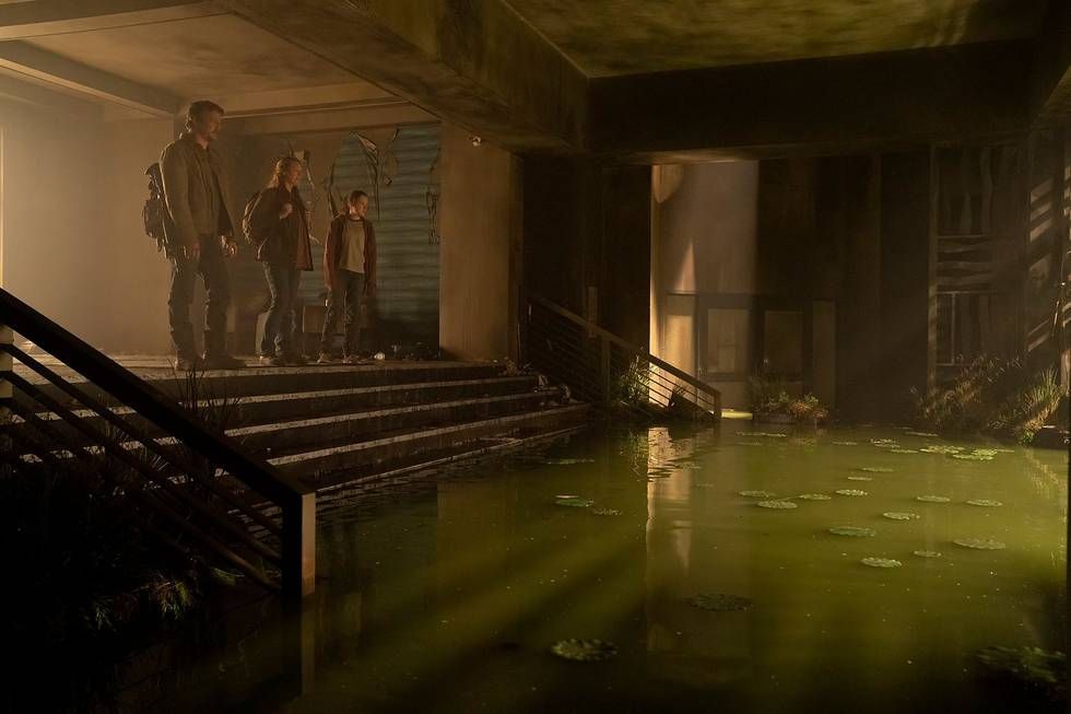 Joel (Pedro Pascal), Tess (Anna Torv), and Ellie (Bella Ramsey) stand at the top of a small staircase of a hotel lobby that has been flooded