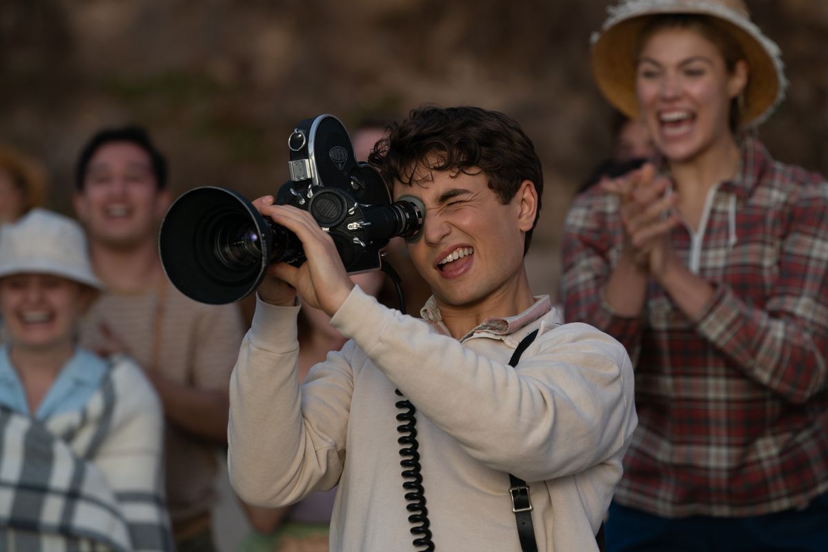 Teenage Sammy Fabelman (Gabriel LaBelle) grins as he points a large film camera at something offscreen while adults behind him smile and cheer in The Fabelmans
