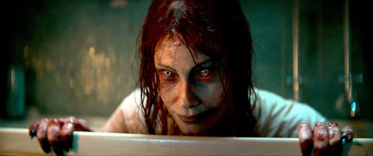 Ellie (Alyssa Sutherland), bezeten en veranderd in een Deadite met rode en gele ogen, vettig haar en een groezelig gezicht, glimlacht griezelig over een barrière in Evil Dead Rise