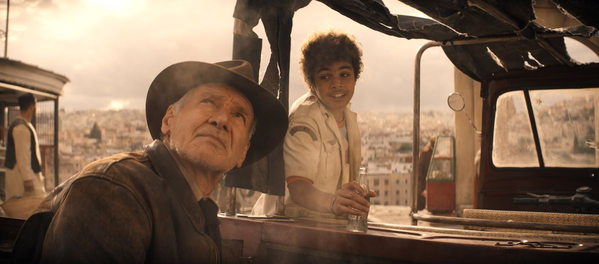 Indiana Jones (Harrison Ford) kijkt omhoog naar een bewolkte grijze lucht terwijl een grijnzende tiener (Ethann Isidore) met hem probeert te praten terwijl hij zich klaarmaakt voor een ritje in zijn vrachtwagen in Indiana Jones and the Dial of Destiny