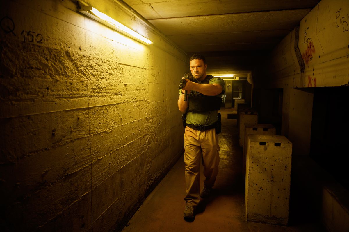Alban Lenoir walks through a long hallway while holding an assault rifle in AKA.