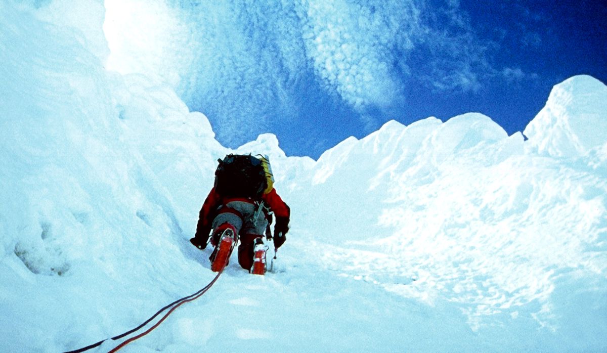 Un alpinista, fotografiado desde abajo, escala una cara de hielo puro, con cielo azul y nubes arriba