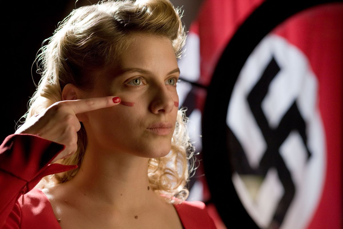Una joven rubia con un vestido rojo se unta pintura de guerra en la cara con una esvástica nazi colgando en el fondo