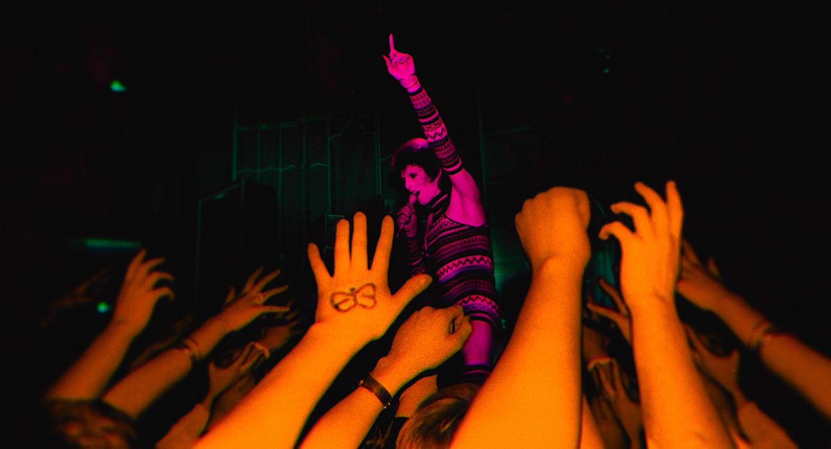 ديفيد بوي ، المظلل باللون الوردي ، في ذروة السبعينيات من القرن الماضي ، يغني ويشير إلى السماء. في المقدمة ، تمد يد المعجبين نحوه